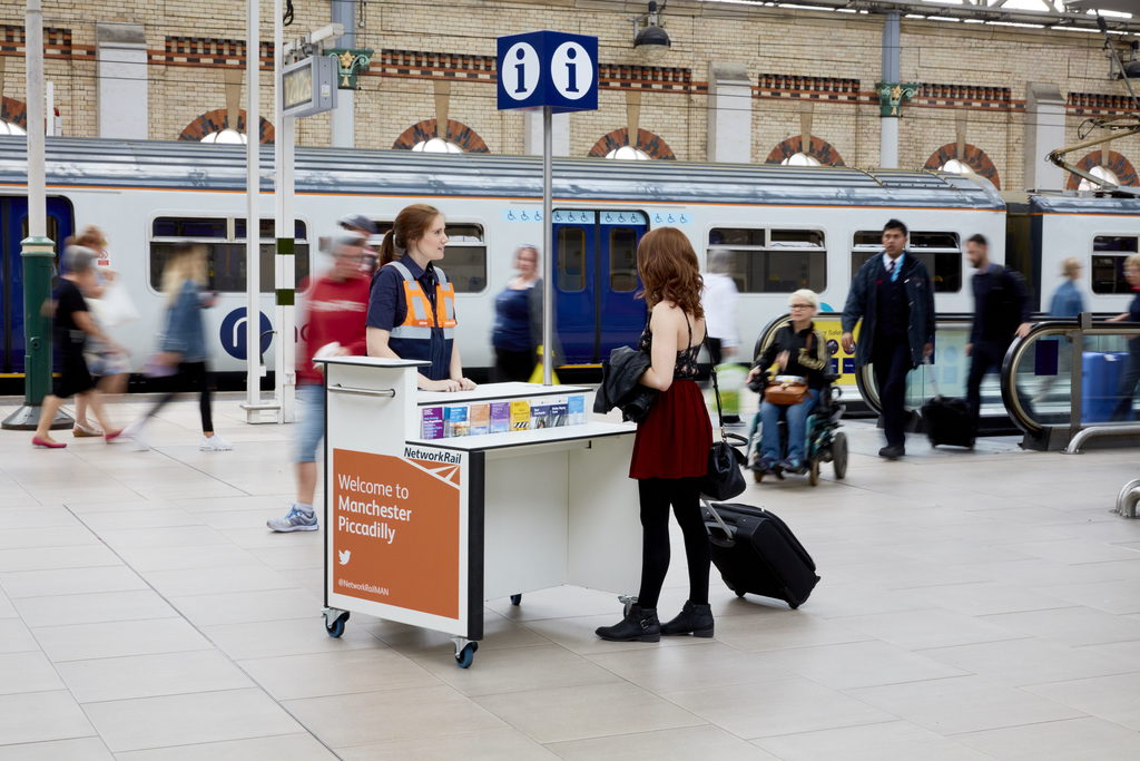 A busy Manchester Piccadilly station concourse with a staffed Network Rail mobile information desk assisting a traveller, surrounded by passengers, clear information signage, and active platform access, illustrating Ottetto’s focus on inclusive passenger assistance, equitable wayfinding, and accessible rail‑station environments.