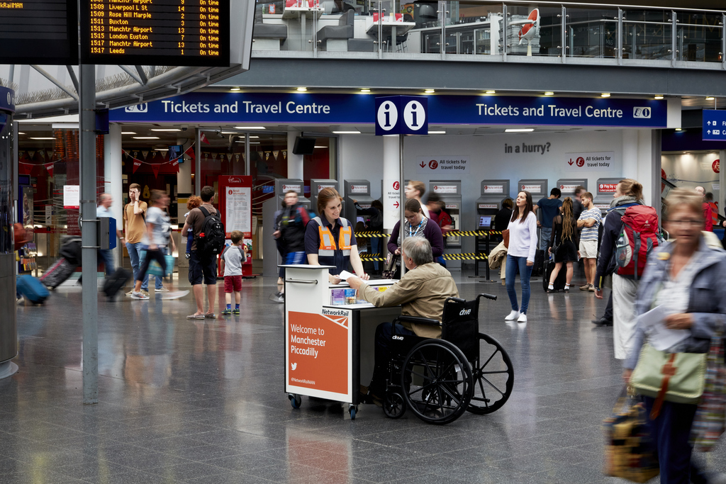 A busy Manchester Piccadilly concourse where a Network Rail staff member supports a wheelchair user at a mobile information desk, reflecting Ottetto’s commitment to equitable passenger assistance, accessible wayfinding, and inclusive transport‑environment design.
